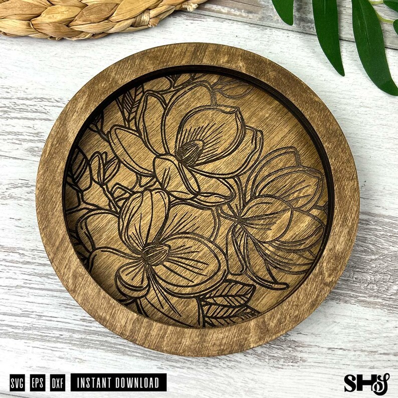 May include: A circular wooden coaster featuring a laser-engraved floral design of magnolia blossoms with detailed petals and leaves. The natural wood grain is visible throughout the piece, which has a raised outer rim and a recessed center.