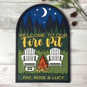 May include: An arched, layered wooden wall sign featuring a night scene with a crescent moon, stars, and a forest silhouette. The text reads "WELCOME TO OUR Fire Pit" and "PAT, ROSE & LUCY" above two white Adirondack chairs, a campfire, and a dog bowl.