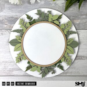 May include: A circular layered wall sign featuring a white center disc framed by a light brown wooden hoop. The perimeter is decorated with various die-cut paper leaves and sprigs in multiple shades of green, including sage and olive tones.