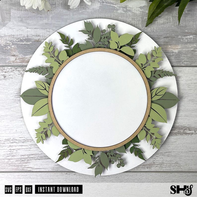 May include: A circular layered wall sign featuring a white center disc framed by a light brown wooden hoop. The perimeter is decorated with various die-cut paper leaves and sprigs in multiple shades of green, including sage and olive tones.