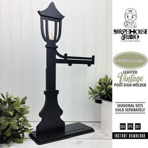 Op de afbeelding: Een zwarte, lasergesneden houten lantaarnpaal-bordhouder met een lantaarn inclusief klein lampje, een horizontale arm voor het ophangen van borden en een rechthoekige voet. De tekst luidt SHAPE HOUSE STUDIO, INTERCHANGEABLE LIGHTED VINTAGE POST SIGN HOLDER, SEASONAL SETS SOLD SEPARATELY, SVG EPS DXF, INSTANT DOWNLOAD.