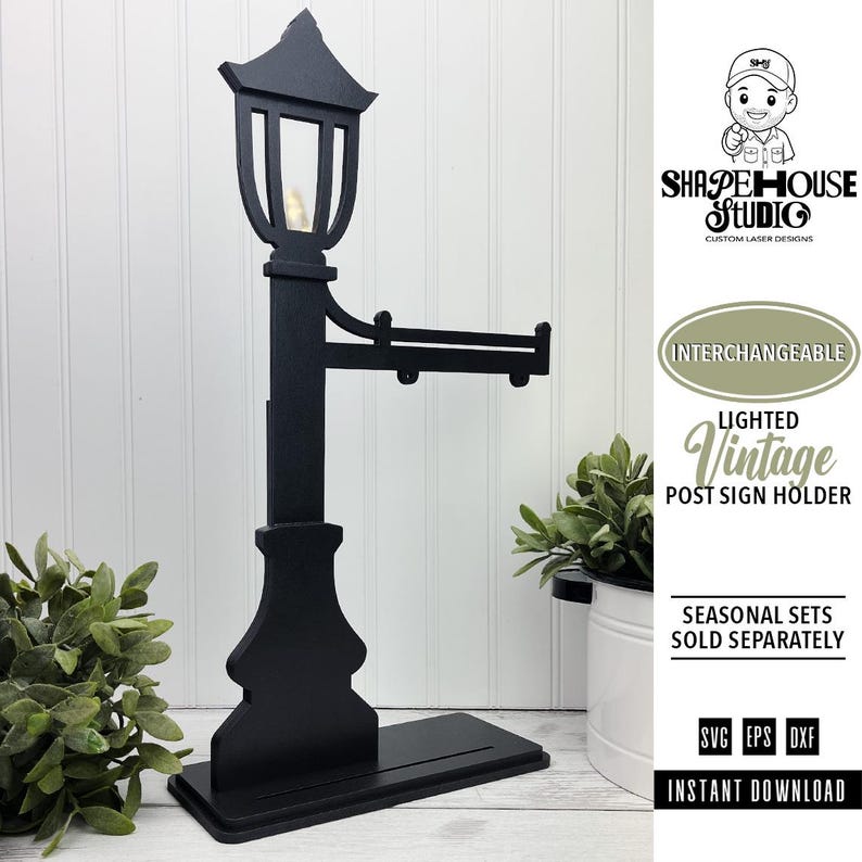 Op de afbeelding: Een zwarte, lasergesneden houten lantaarnpaal-bordhouder met een lantaarn inclusief klein lampje, een horizontale arm voor het ophangen van borden en een rechthoekige voet. De tekst luidt SHAPE HOUSE STUDIO, INTERCHANGEABLE LIGHTED VINTAGE POST SIGN HOLDER, SEASONAL SETS SOLD SEPARATELY, SVG EPS DXF, INSTANT DOWNLOAD.