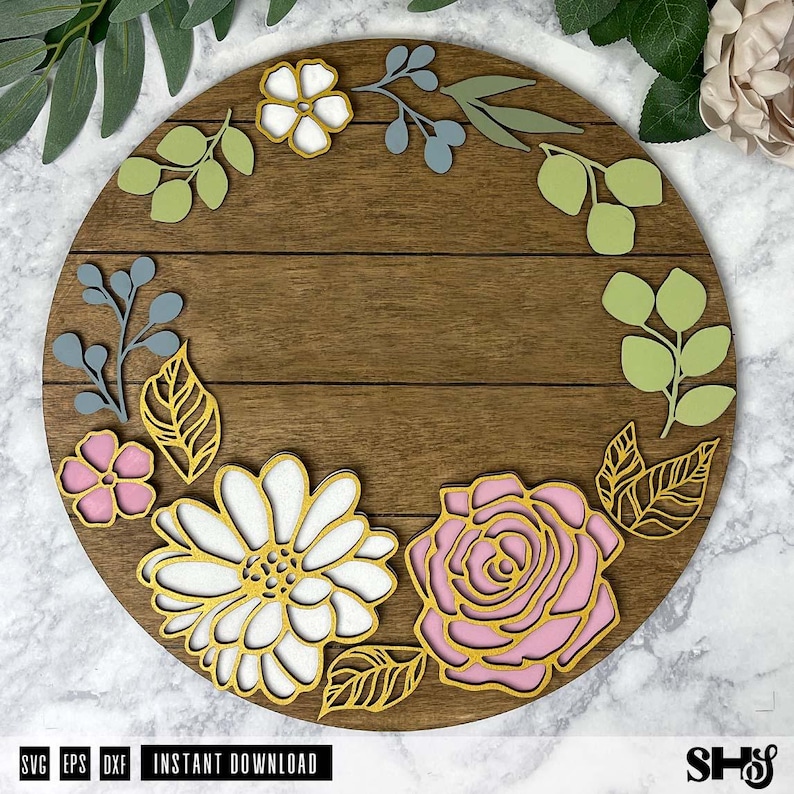 May include: A circular wooden sign featuring a dark brown horizontal plank design decorated with layered floral and leaf cutouts in shades of white, light pink, sage green, and muted blue, with gold-toned outlines on the flower petals.