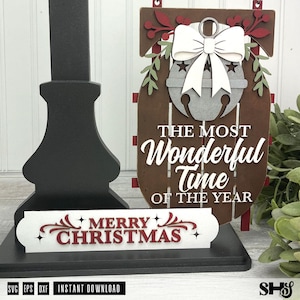 May include: A brown wooden sled-shaped sign featuring a grey jingle bell with a white bow, green leaves, and red berries. The text reads THE MOST WONDERFUL TIME OF THE YEAR. A separate white rectangular sign below reads MERRY CHRISTMAS in red.