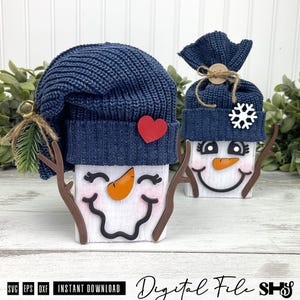 May include: Two wooden snowman figures featuring white painted blocks with black smiling faces, orange carrot noses, and pink cheeks. Each wears a dark blue knit hat. One hat has a red heart, the other a white snowflake and twine bow.