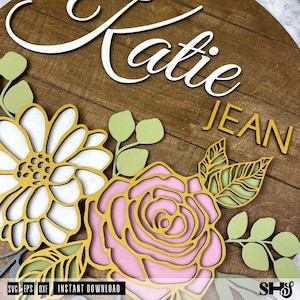 Op de afbeelding: Een houten bord met de namen "Katie" en "Jean" in witte letters. Het bord is versierd met een bloemmotief met roze, witte en groene bloemen.