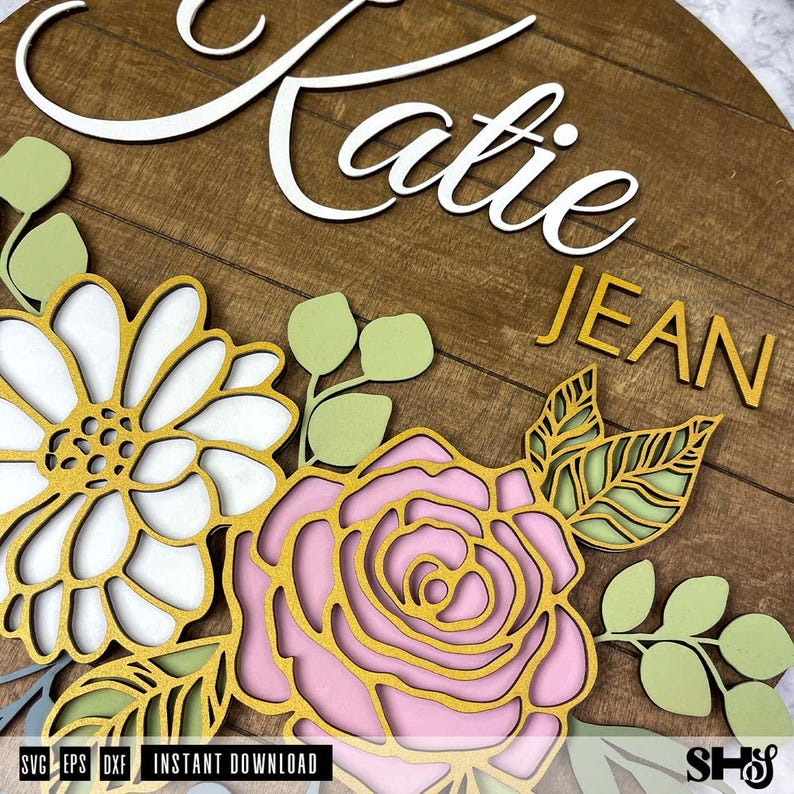 Op de afbeelding: Een houten bord met de namen "Katie" en "Jean" in witte letters. Het bord is versierd met een bloemmotief met roze, witte en groene bloemen.