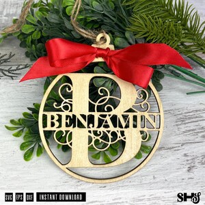 Op de afbeelding: Een rond houten ornament met een grote uitgesneden letter B en de naam BENJAMIN in het midden verwerkt. Het ornament is versierd met een felrood satijnen lint in een strik aan de bovenkant en heeft een jute touwtje om op te hangen.
