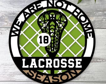 Może przedstawiać: Okrągła tabliczka ścienna z czarną sylwetką kija do lacrosse umieszczoną na limonkowym tle z białym wzorem kratki. Tekst WE ARE NOT HOME oraz LACROSSE SEASON otacza grafikę, a liczba 18 znajduje się wewnątrz białego koła.