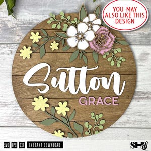 May include: A circular wooden sign featuring the name Sutton in large white script and Grace in smaller pink block letters. The design is decorated with layered wooden floral elements in white, soft pink, and sage green, including small yellow flowers.