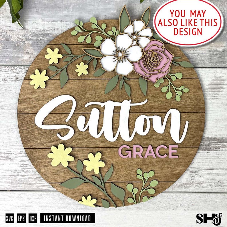 May include: A circular wooden sign featuring the name Sutton in large white script and Grace in smaller pink block letters. The design is decorated with layered wooden floral elements in white, soft pink, and sage green, including small yellow flowers.