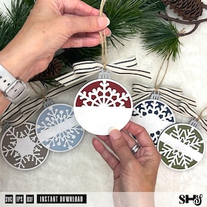 May include: A collection of round, flat, wooden Christmas ornaments featuring intricate snowflake cutouts. Each ornament has a solid horizontal band across the centre for personalisation, a striped fabric ribbon bow, and a jute twine hanging loop.