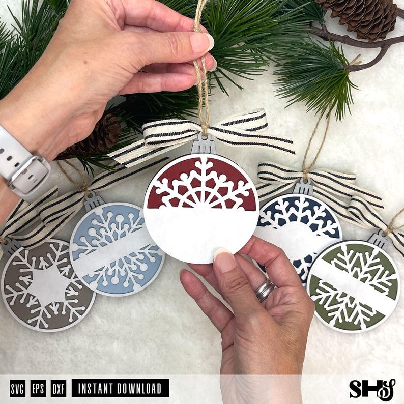 May include: A collection of round, flat, wooden Christmas ornaments featuring intricate snowflake cutouts. Each ornament has a solid horizontal band across the centre for personalisation, a striped fabric ribbon bow, and a jute twine hanging loop.