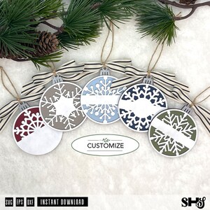 May include: Five circular ornaments featuring intricate snowflake cutouts in maroon, beige, light blue, dark blue, and olive green. Each ornament has a white horizontal banner for personalisation, a striped fabric bow, and a jute twine hanger.