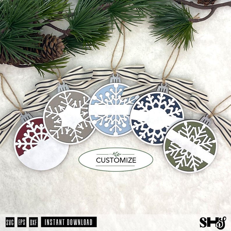 May include: Five circular ornaments featuring intricate snowflake cutouts in maroon, beige, light blue, dark blue, and olive green. Each ornament has a white horizontal banner for personalisation, a striped fabric bow, and a jute twine hanger.