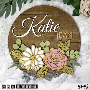 Op de afbeelding: Een houten bord met de namen "Katie Jean" in witte letters op een bruine achtergrond. Het bord is versierd met een bloemmotief met witte, roze en gele bloemen.