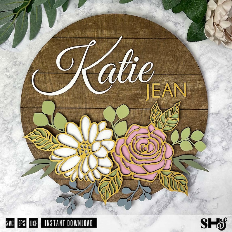 Op de afbeelding: Een houten bord met de namen "Katie Jean" in witte letters op een bruine achtergrond. Het bord is versierd met een bloemmotief met witte, roze en gele bloemen.