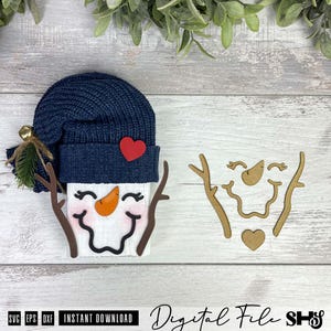 May include: A snowman craft project featuring a navy blue knit hat with a small gold bell, a red heart accent, and a white face with a carrot nose and branch arms. Beside it are separate wooden cutouts of the snowman face, arms, and heart.
