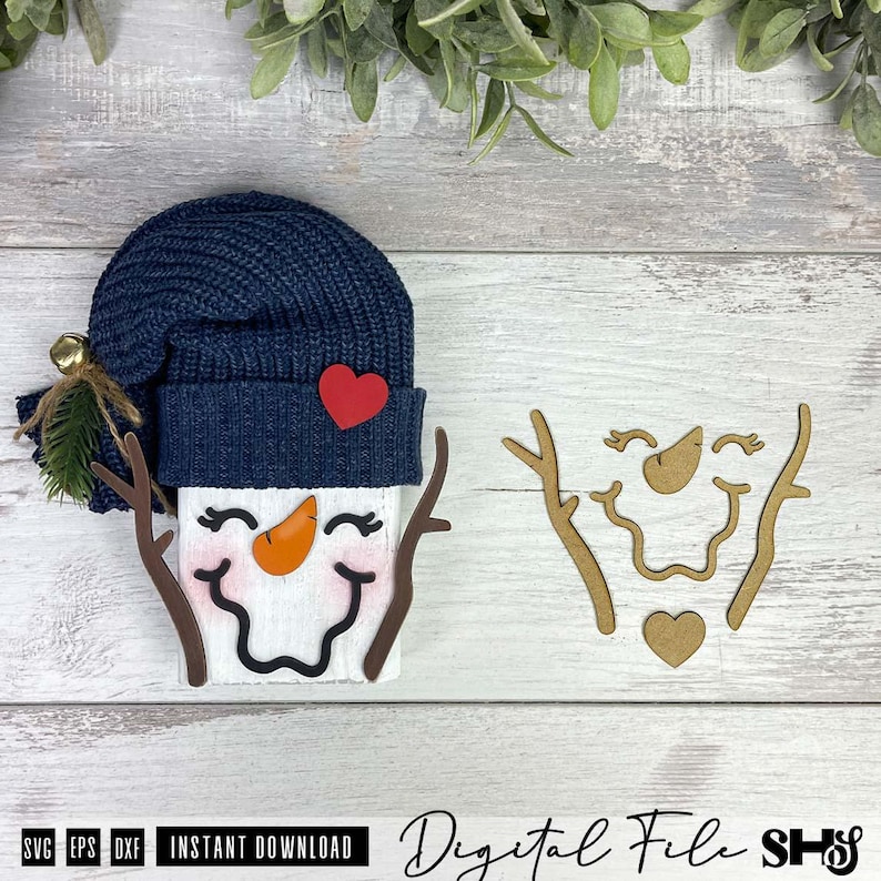 May include: A snowman craft project featuring a navy blue knit hat with a small gold bell, a red heart accent, and a white face with a carrot nose and branch arms. Beside it are separate wooden cutouts of the snowman face, arms, and heart.