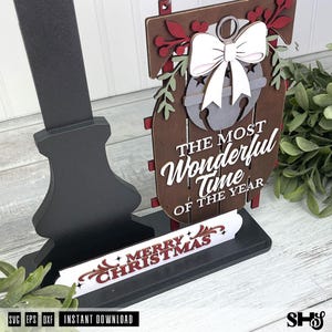 May include: A layered wooden sign featuring a dark brown sled shape with a white bow and bell cutout. The text reads THE MOST WONDERFUL TIME OF THE YEAR. A separate white base piece displays the red text MERRY CHRISTMAS. The sign is mounted on a black stand.