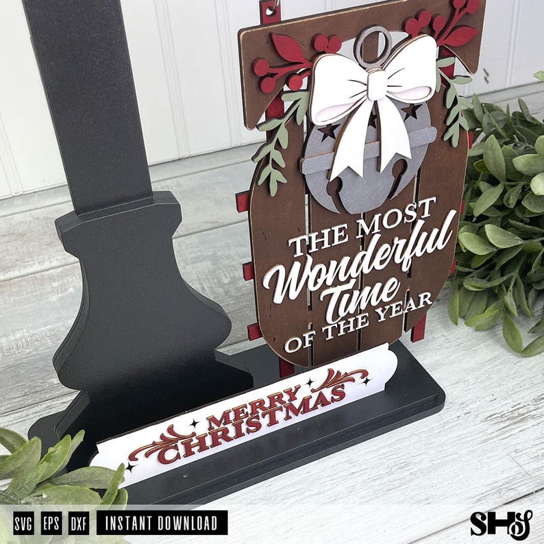 May include: A layered wooden sign featuring a dark brown sled shape with a white bow and bell cutout. The text reads THE MOST WONDERFUL TIME OF THE YEAR. A separate white base piece displays the red text MERRY CHRISTMAS. The sign is mounted on a black stand.