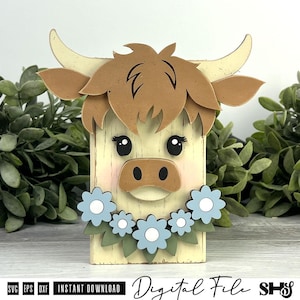 Pu&ograve; includere: Una sagoma in legno a forma di mucca Highland con pelliccia color crema, capelli castani e corna ricurve. La mucca presenta occhi neri, guance rosa e un muso marrone. Una ghirlanda di fiori azzurri con foglie verdi si trova sotto.