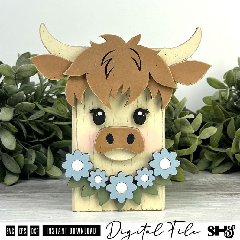 Pu&ograve; includere: Una sagoma in legno a forma di mucca Highland con pelliccia color crema, capelli castani e corna ricurve. La mucca presenta occhi neri, guance rosa e un muso marrone. Una ghirlanda di fiori azzurri con foglie verdi si trova sotto.