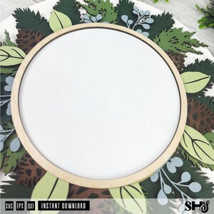 May include: A circular wooden frame centered over a layered botanical design featuring dark green pine needles, light green leaves, brown pinecones, and pale blue sprigs. The design is flat and multi-layered with a clean white center space.