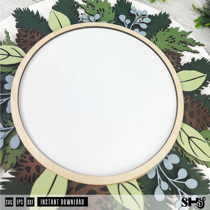 May include: A circular wooden frame centered over a layered botanical design featuring dark green pine needles, light green leaves, brown pinecones, and pale blue sprigs. The design is flat and multi-layered with a clean white center space.