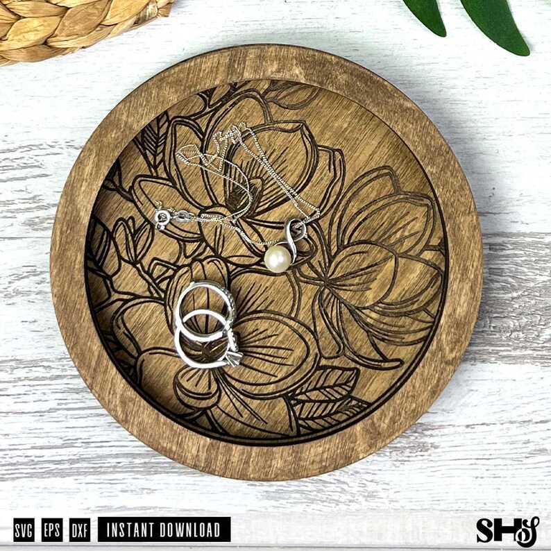 May include: A round wooden tray featuring a laser-engraved floral pattern on its base. Resting inside the tray are a silver-toned necklace with a single white pearl pendant and two silver-toned rings stacked together on the engraved surface.