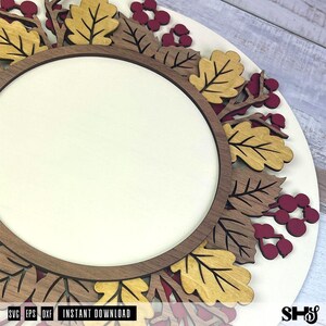 May include: A circular wooden sign featuring a central blank cream-colored disc framed by a dark brown ring. The border is decorated with layered laser-cut oak leaves in shades of mustard yellow and brown, interspersed with small dark red berries.