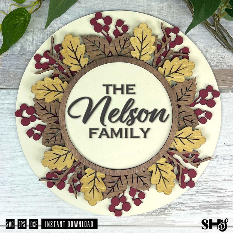 May include: A circular wooden sign featuring a layered design of oak leaves in shades of gold and brown, interspersed with dark red berries. The center circle displays the text THE Nelson FAMILY in a combination of block and script lettering.