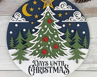 May include: A layered wooden Christmas countdown decoration. The design features a Christmas tree with red ornaments, a crescent moon, stars, and the text "DAYS UNTIL CHRISTMAS". The top star displays the number 25.