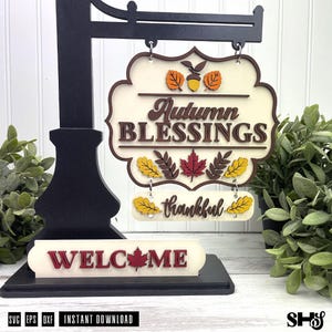 Op de afbeelding: Een zwarte metalen standaard met een gelaagd houten bord met de tekst Autumn BLESSINGS en thankful. Het bord bevat herfstbladuitsnijdingen in oranje, geel en rood. Een apart basisstuk toont het woord WELCOME met een rood esdoornblad.