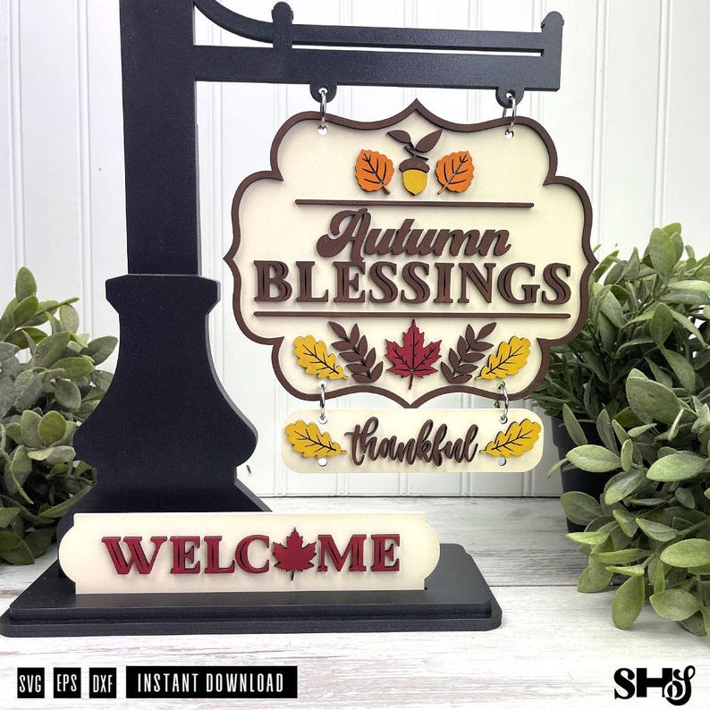 Op de afbeelding: Een zwarte metalen standaard met een gelaagd houten bord met de tekst Autumn BLESSINGS en thankful. Het bord bevat herfstbladuitsnijdingen in oranje, geel en rood. Een apart basisstuk toont het woord WELCOME met een rood esdoornblad.