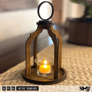 May include: A dark brown wooden lantern with an arched frame and a circular top handle. The lantern features a clear glass votive holder containing a small, glowing orange LED tea light candle. The base is circular with etched floral details.