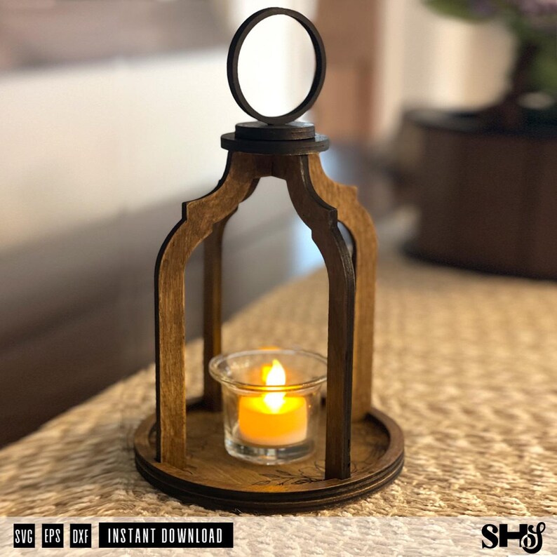 May include: A dark brown wooden lantern with an arched frame and a circular top handle. The lantern features a clear glass votive holder containing a small, glowing orange LED tea light candle. The base is circular with etched floral details.