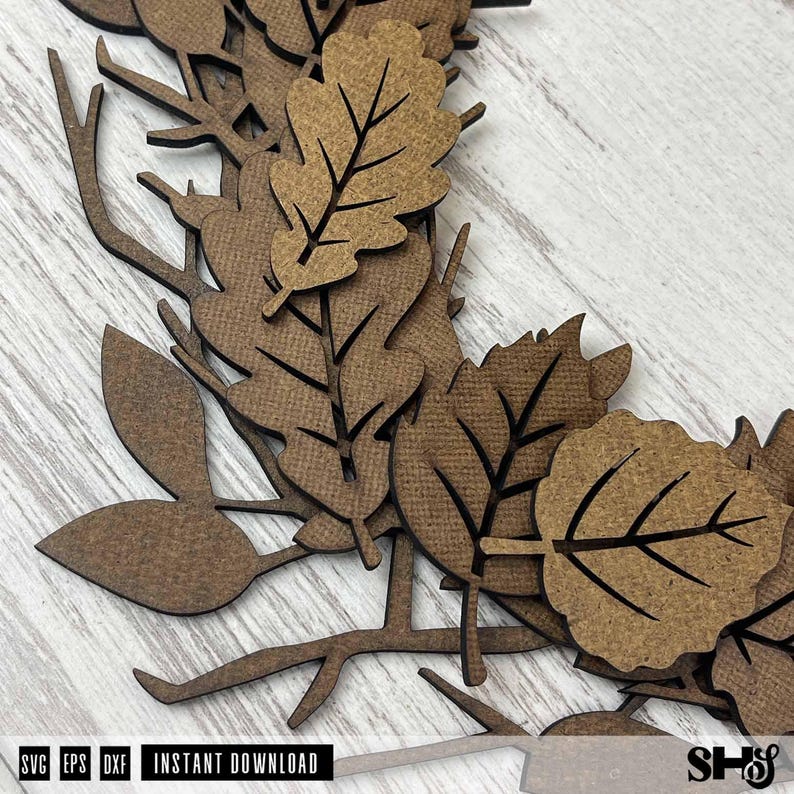 Puede incluir: Una decoraci&oacute;n de corona de madera marr&oacute;n cortada con l&aacute;ser que presenta hojas de roble y abedul superpuestas con detalles de venas intrincados y ramas delgadas. El borde inferior incluye el texto SVG, EPS, DXF, INSTANT DOWNLOAD y SHS.
