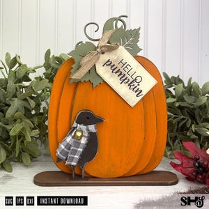 May include: A decorative orange pumpkin cutout featuring a green leaf, a burlap ribbon, and a tag with the text "HELLO pumpkin". A black bird figure with a black and white plaid scarf and a small gold bell is attached to the front of the pumpkin.