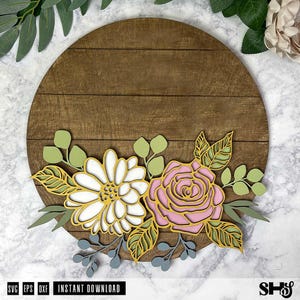Op de afbeelding: Een rond houten bord met een bloemmotief. Het ontwerp bevat een roze roos, een witte madeliefje en groene bladeren. Het bord is perfect voor een boerderij- of rustieke inrichting.