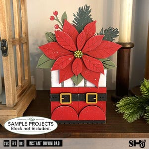 May include: A decorative layered cutout featuring a red poinsettia flower with green leaves and pine sprigs atop a white block with a black and white striped pattern and a red base resembling boots with a black belt and gold buckles.