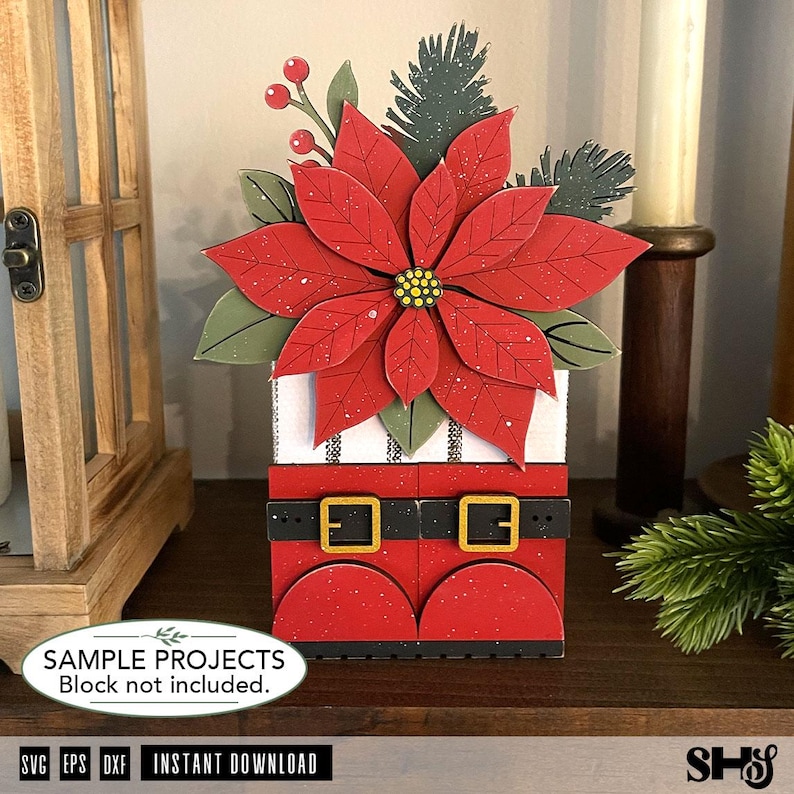 May include: A decorative layered cutout featuring a red poinsettia flower with green leaves and pine sprigs atop a white block with a black and white striped pattern and a red base resembling boots with a black belt and gold buckles.