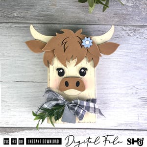 Pu&ograve; includere: Una sagoma in legno a forma di testa di mucca highland con pelo color crema, capelli marroni e corna ricurve. Presenta un piccolo fiore azzurro, occhi neri, un naso marrone e un nastro a quadri grigio e bianco legato con foglie verdi.