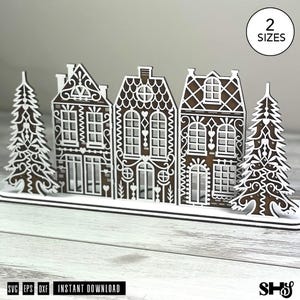 May include: A laser-cut wooden gingerbread village display featuring three houses and two pine trees with white decorative icing-style patterns. The design includes a base and a circular badge in the top right corner with the text 2 SIZES.