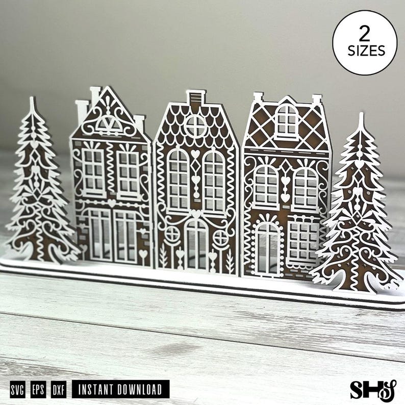 May include: A laser-cut wooden gingerbread village display featuring three houses and two pine trees with white decorative icing-style patterns. The design includes a base and a circular badge in the top right corner with the text 2 SIZES.