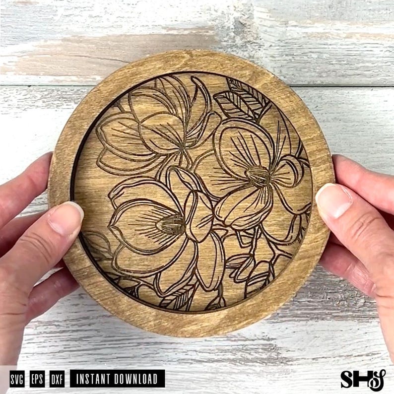 May include: A round wooden coaster featuring a laser-engraved floral design of three large blossoms with detailed petals and leaves. The natural wood grain is visible throughout the circular frame, and the engraved lines are dark brown.