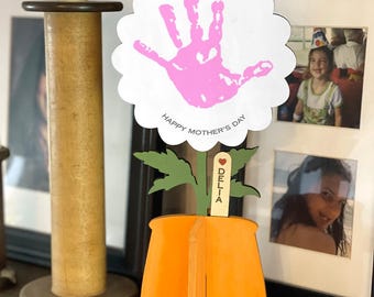 May include: A wooden craft project for Mother's Day. A white flower with a pink handprint in the center. The flower is attached to a green stem and a brown pot. The pot has a wooden sign that says "Happy Mother's Day" and a smaller sign that says "Delia".