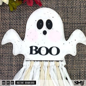 May include: A white ghost-shaped wooden cutout with black oval eyes, a small round mouth, and pink cheeks. The word BOO is printed in black letters across the center. Below the ghost, a fringe of white and beige fabric strips hangs down.