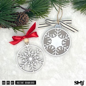 May include: Two circular snowflake ornaments. The left ornament is white with a red satin ribbon and a wooden bead. The right ornament is grey with a white snowflake cutout, topped with a black and white striped ribbon and a jute twine loop.