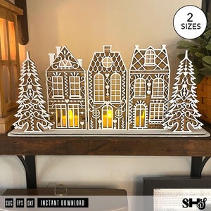 May include: A laser-cut wooden gingerbread village display featuring three houses and two pine trees with intricate white filigree detailing. The structures are arranged on a flat base, with yellow LED tea lights glowing inside the houses.